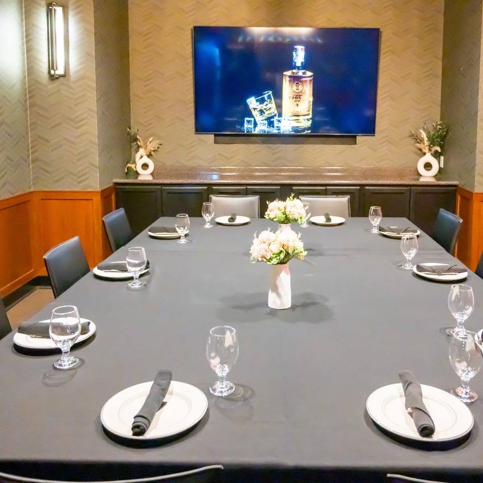 A private dining room with a long table set with plates, glasses, and floral centerpieces, under a large TV.