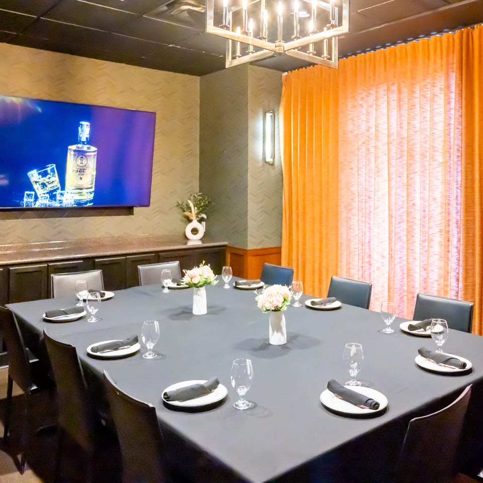 A private dining room with a large square table set for guests, a TV, and orange curtains.