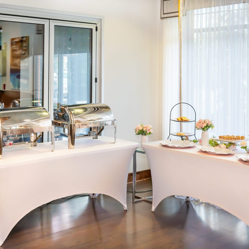 Elegant buffet setup with covered chafing dishes, pastries, floral arrangements, and white-draped tables beside bright windows.