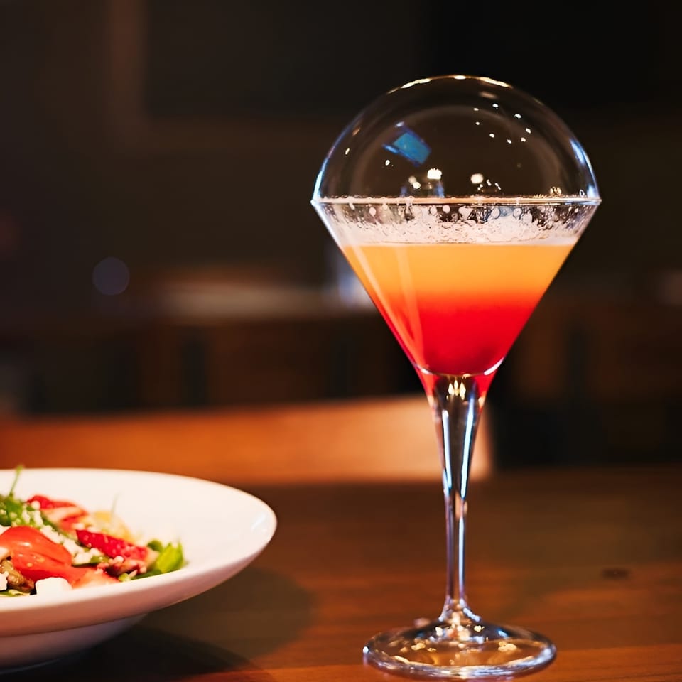A vibrant red and orange cocktail with a bubble on top, next to a fresh salad.