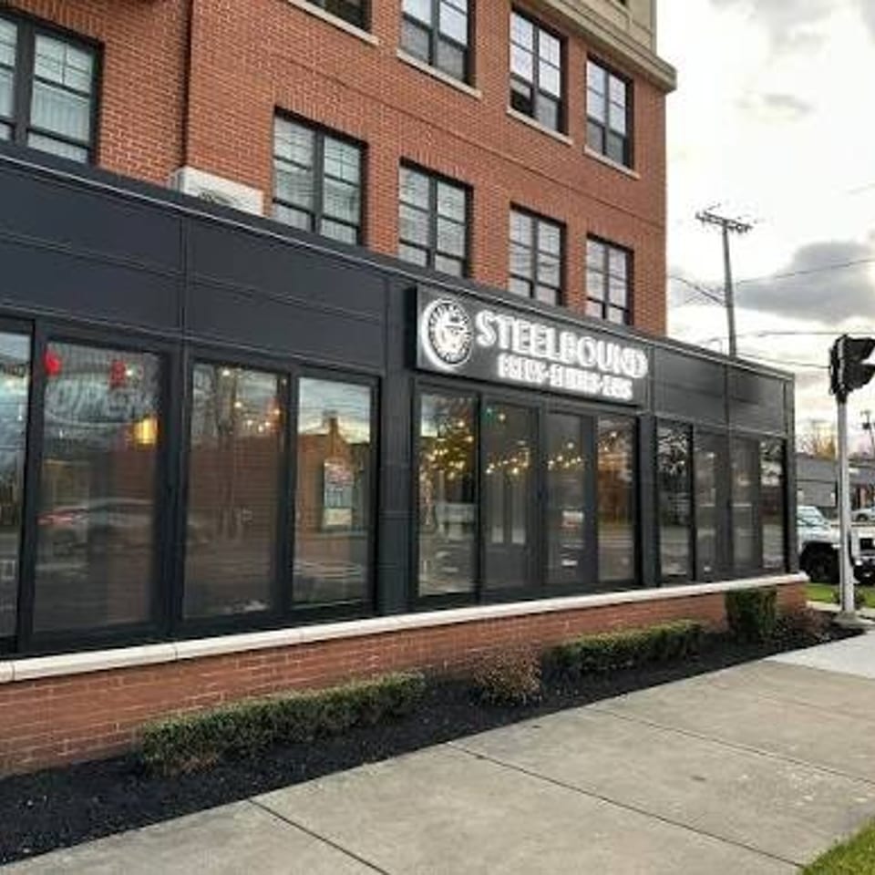 Brick restaurant storefront with dark-framed windows, sidewalk landscaping, and an illuminated Steel... sign beneath an overcast sky.