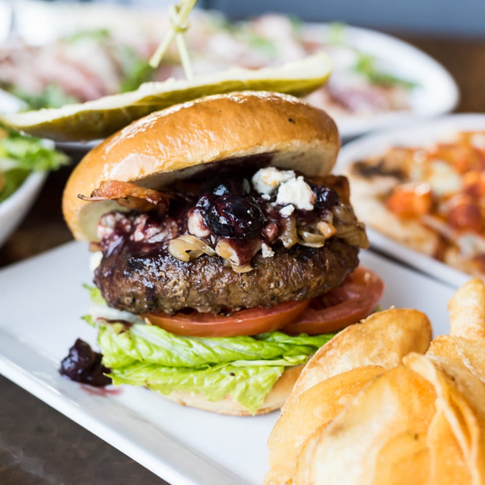 Gourmet burger topped with bacon, blue cheese, and berry compote, served with chips.