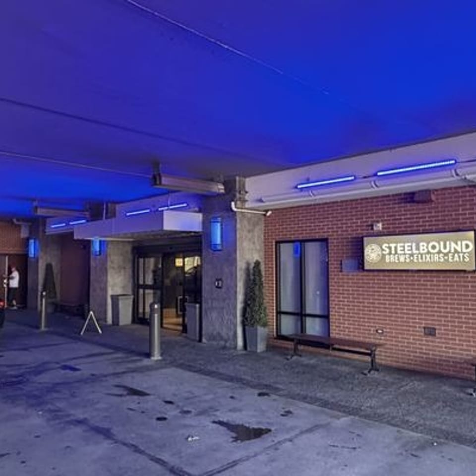 Blue-lit covered restaurant entrance with brick walls, benches, and a Steel Bound Brews-Elixirs-Eats sign beside a parked car.