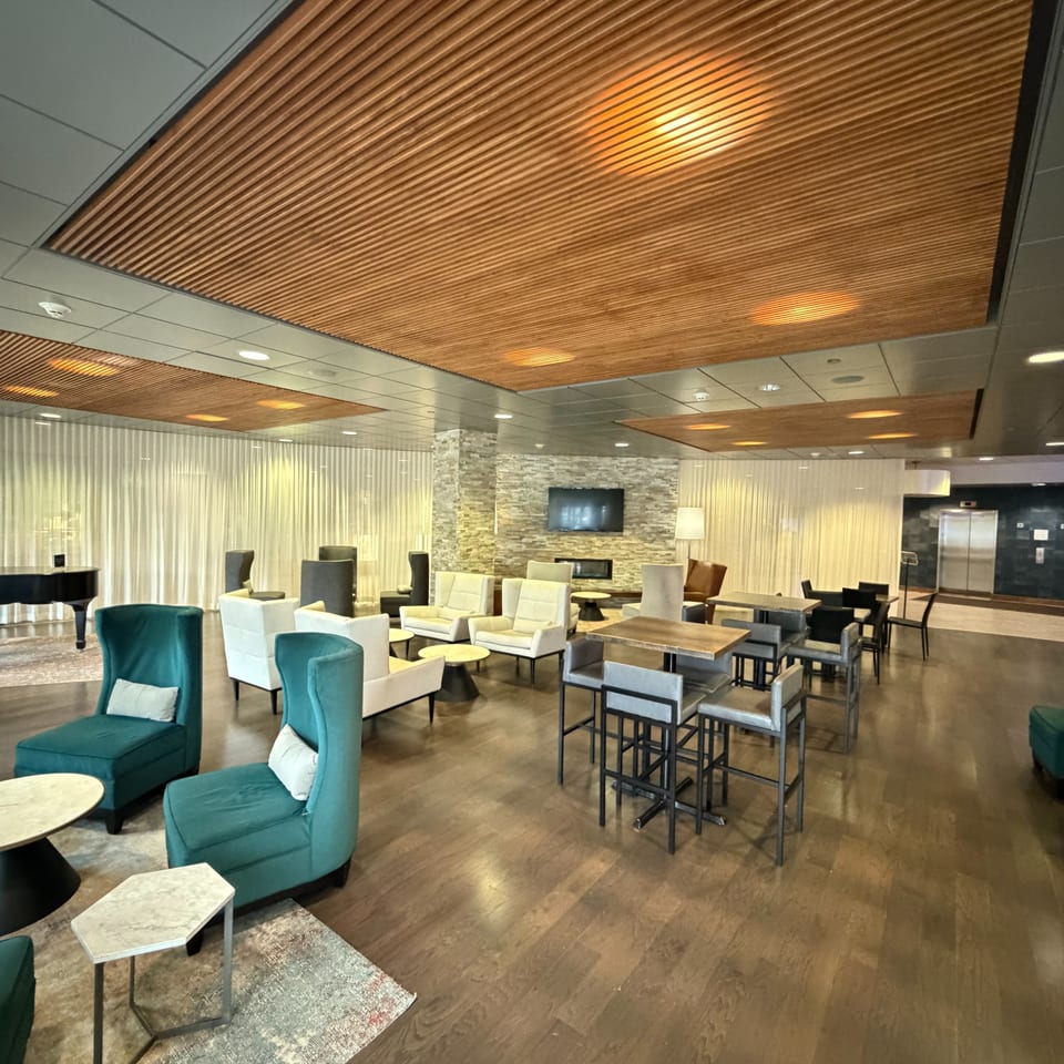 Modern lounge with teal and cream seating, marble tables, a grand piano, stone fireplace wall, and warm wood ceiling accents.