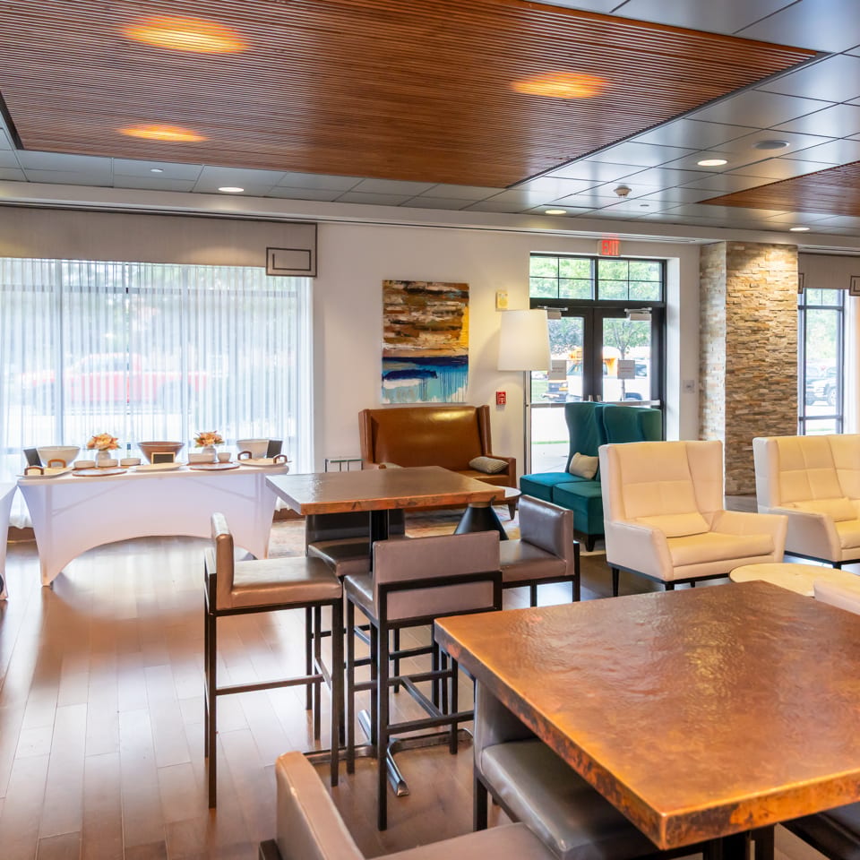 Bright restaurant lounge with modern seating, wood tables, stone columns, buffet setup, artwork, and a grand piano.