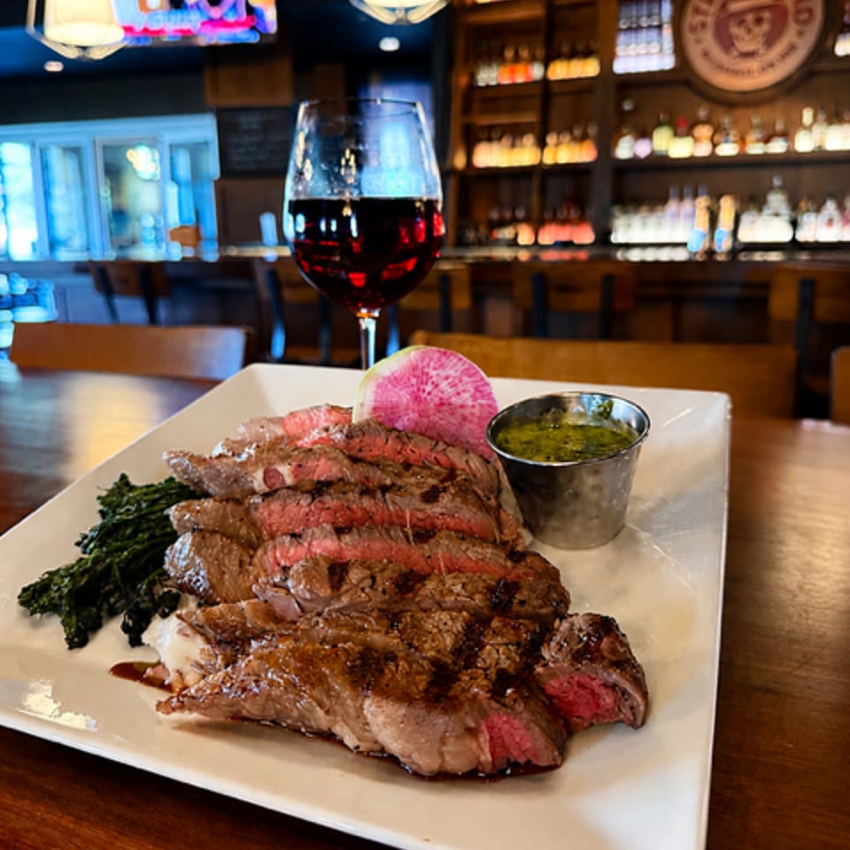 Sliced medium-rare steak with grilled greens, radish, and dipping sauce on a white plate beside red wine in a warmly lit bar.