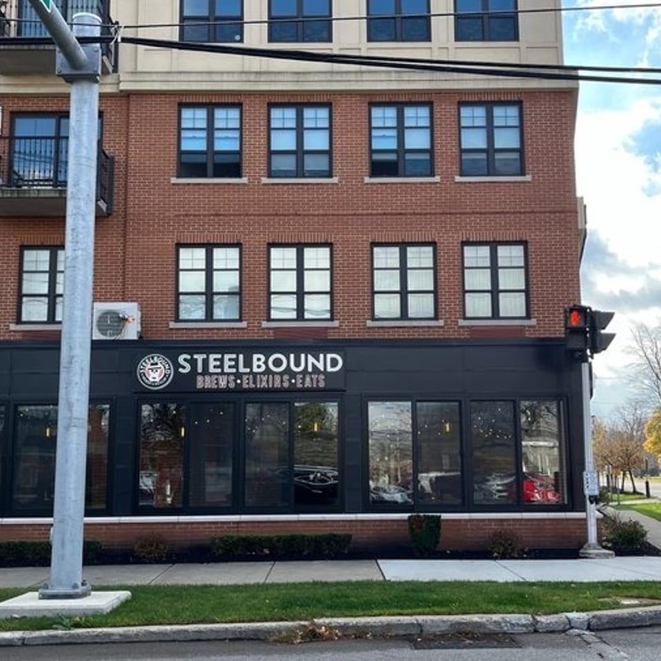 Street view of Steelbound Brewery, showing a brick building, storefront, sidewalk, traffic light, parked cars, and blue sky.