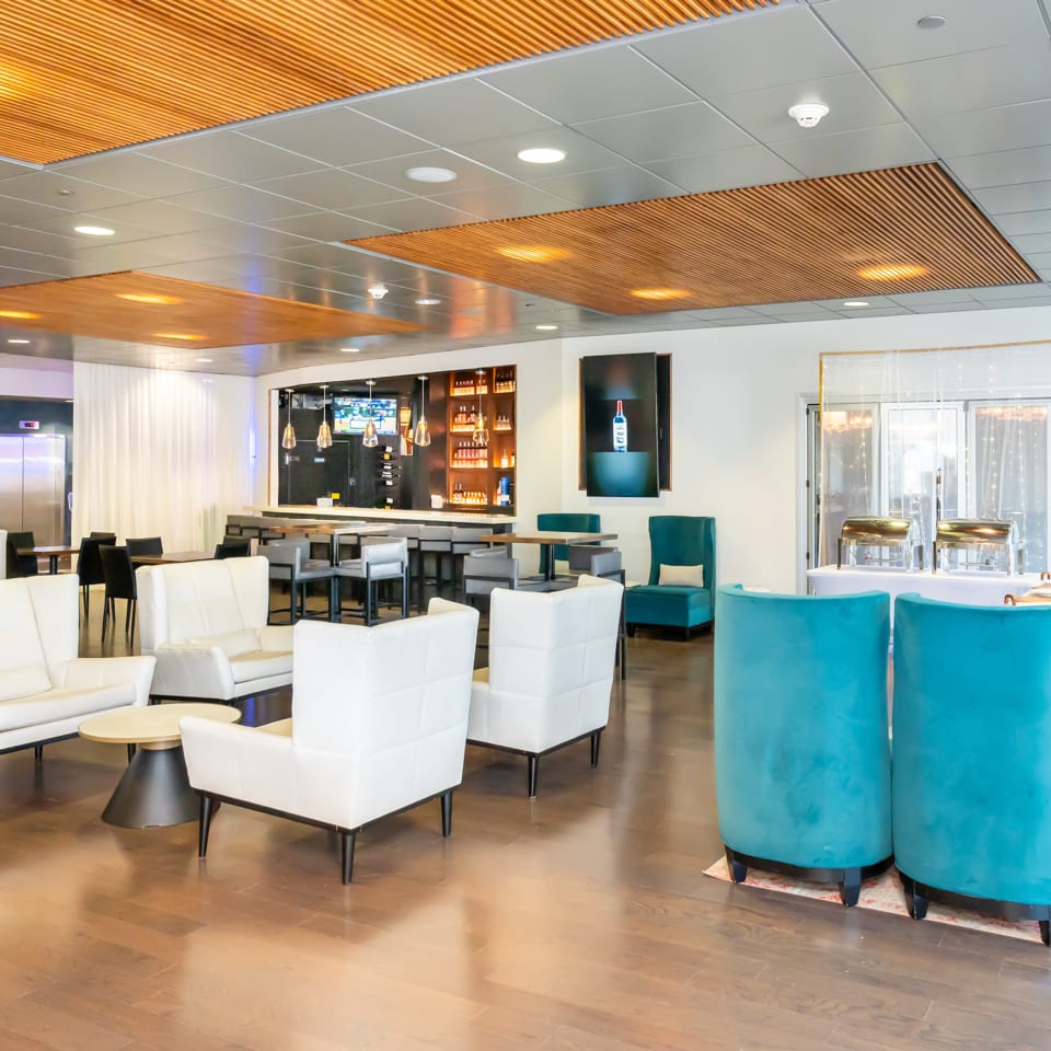 Modern restaurant lounge with white armchairs, turquoise seating, wood-paneled ceiling, bar, and buffet station.