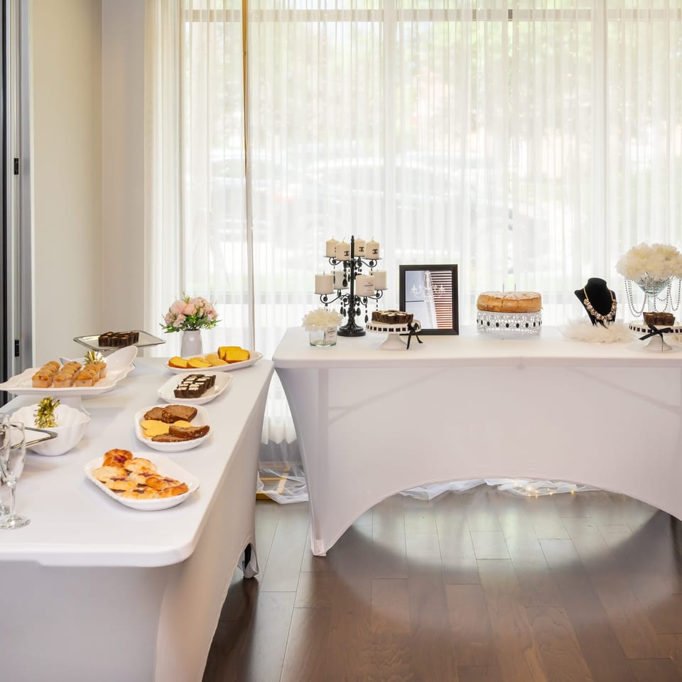 Elegant dessert buffet with pastries, cakes, floral arrangements, jewelry displays, and champagne beneath bright sheer-curtained windows.