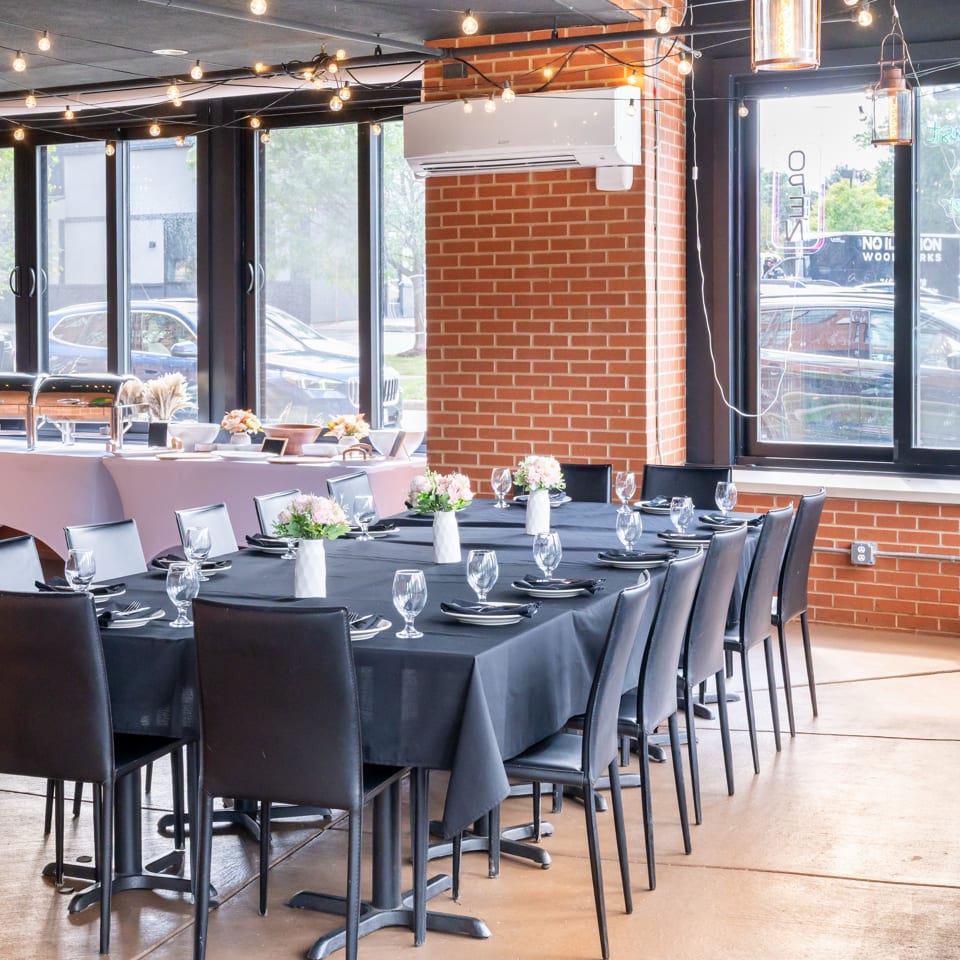 Restaurant dining room with black table settings, floral centerpieces, buffet stations, exposed brick, large windows, and string lights.