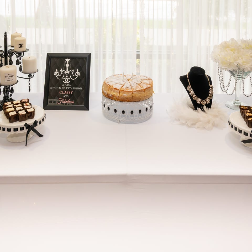 Elegant Chanel-themed dessert table with candles, cake, brownies, jewelry displays, flowers, and a sign reading “A girl should be two things: classy and fabulous.”