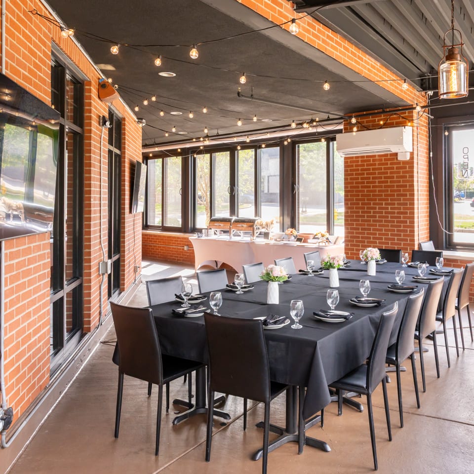 Brick-walled restaurant dining room with black table settings, floral centerpieces, large windows, and warm string lights.