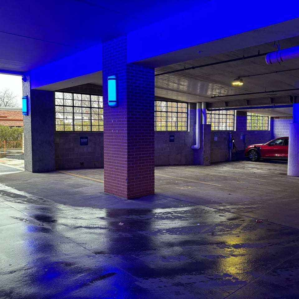 Covered parking garage with blue lighting, wet pavement, brick columns, large windows, and a red car near electric vehicle chargers.