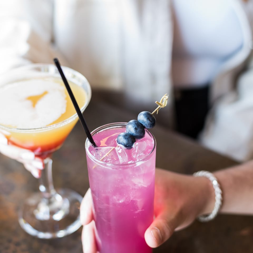 A purple cocktail garnished with blueberries held at the bar next to a citrus martini.