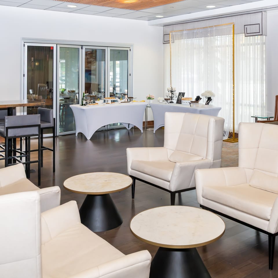 Lounge area with white armchairs, round tables, and a buffet setup in a modern event space.