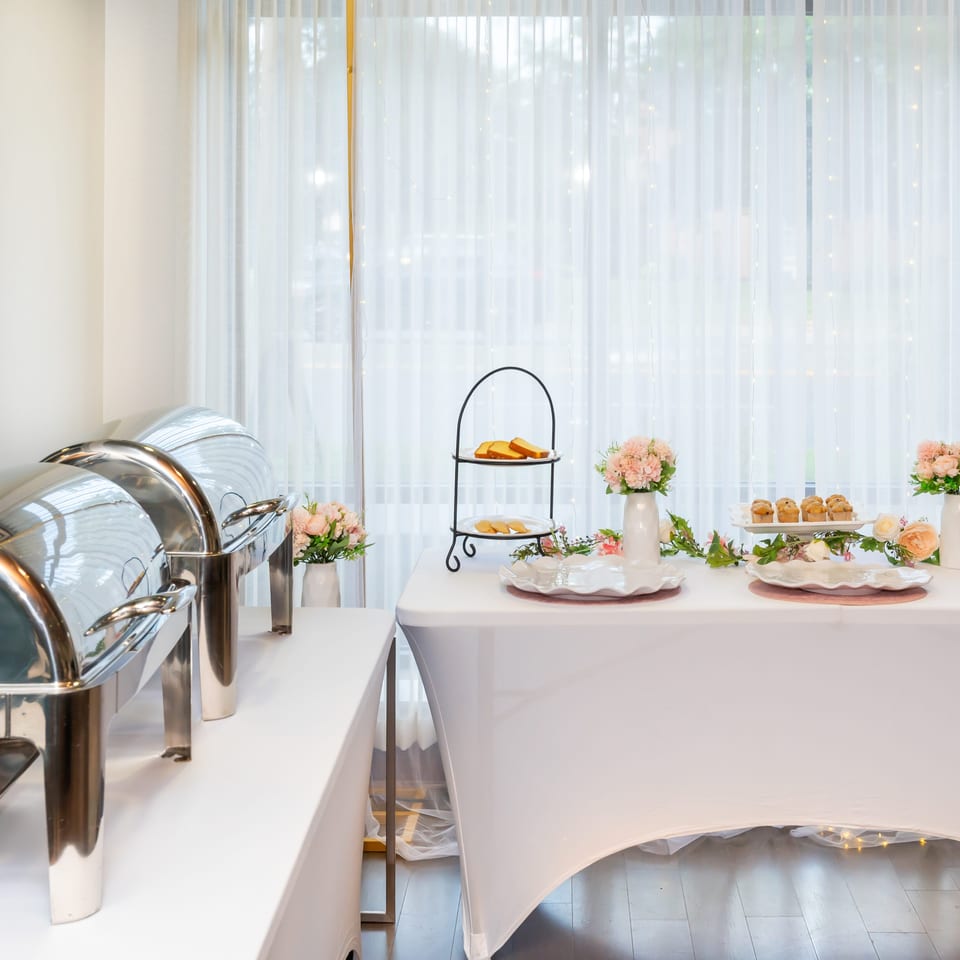 Elegant buffet table with chafing dishes, pastries, tiered stands, and pastel floral arrangements beside sheer windows.